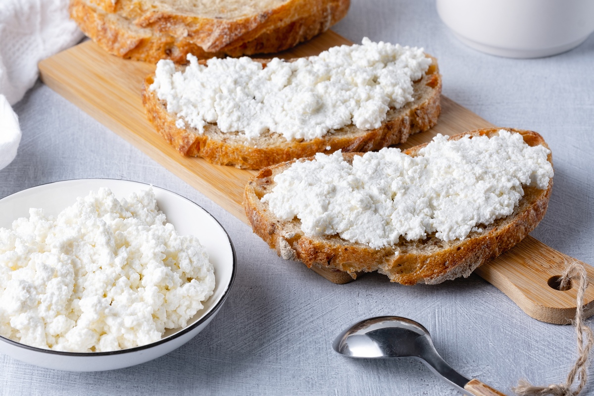 Two slices of bread with curd cheese on wooden board. A bowl of curd cheese and a sliver spoon besides the slices.