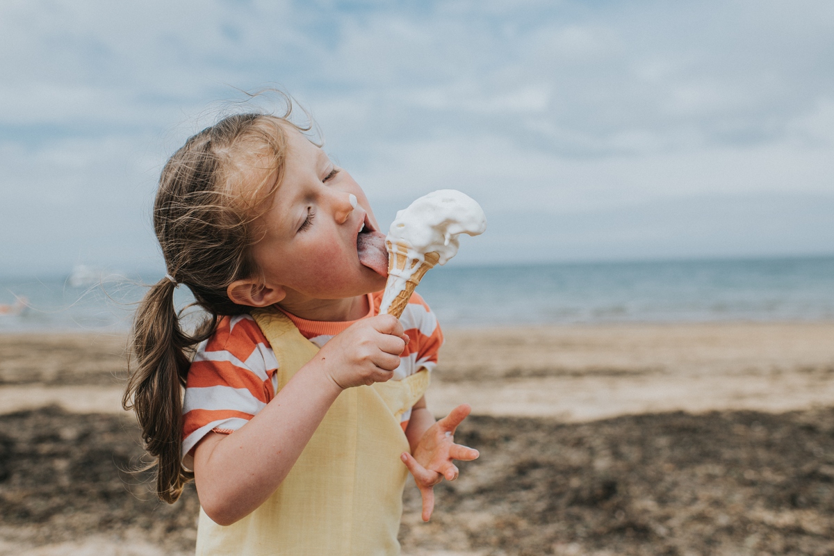 Young girl on the beach, licking an ice cream. Her nose is covered with a drop of ice cream and the waffle in her hand is covered by the molten ice cream. The wind is blowing in her face, pushing back her hair.