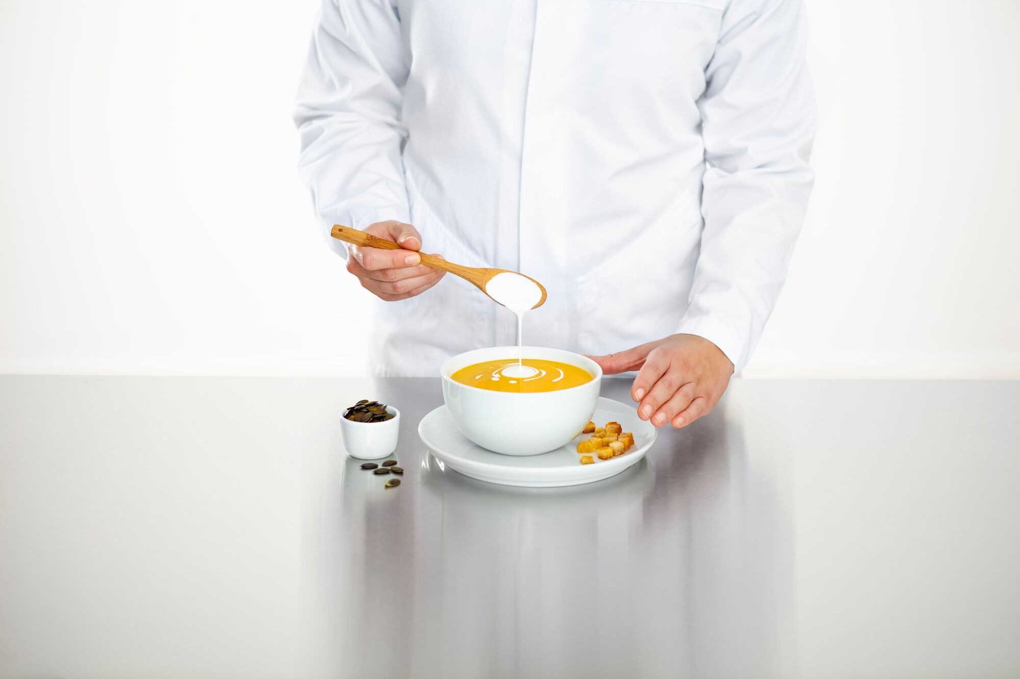 Technologist standing infront of a metallic table, wearing a lab coat, pouring cooking cream into a bowl of pumkin soup.