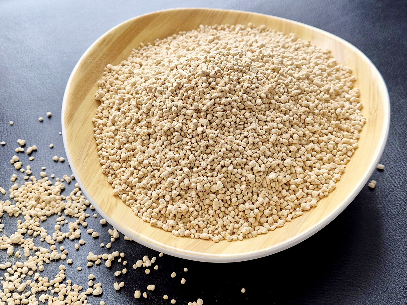 A bowl of lecithin granules in a wooden bowl on a dark table top. Some granules have fallen off and scattered to the side.